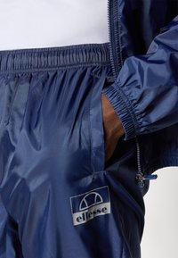 Navy windbreaker en broekset gemaakt van glanzend materiaal. Voorzien van een elastische taille, ritszakken en een wit logo-opnamer op de broek.
