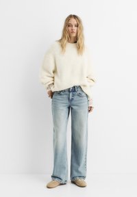 Maglione oversize color crema in maglia texturizzata, abbinato a jeans a gamba larga blu chiaro. L'outfit presenta sottili dettagli in pizzo sulle maniche.