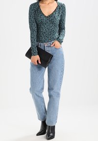 Nainen, joka on pukeutunut teal-väriseen kuvioituun pitkähihaiseen paitaan, vaaleansinisiin korkeavyötäröisiin farkkuihin, mustiin nilkkureihin ja pitää kädessään mustaa laukkuhuppua.