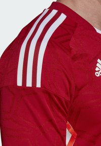 Czerwona sportowa koszulka z teksturowanym wzorem, biała konstrukcja na ramionach w trzy paski, okrągły kołnierz oraz pomarańczowy akcent z boku.