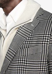 Blazer in bianco e nero con motivo houndstooth, dotato di colletto con zip, caratterizzato da tessuto testurizzato e rever strutturati, indossato sopra una camicia bianca.