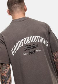T-shirt in cotone marrone con maniche corte e scollatura rotonda, caratterizzata da testo stampato bianco sulla schiena: "GOODFORNOTHING Atelier No.1 Design."