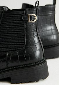 Las botas negras hasta el tobillo presentan un upper de cuero texturizado con un patrón de piel de cocodrilo, un panel de malla y acentos de hardware dorado. Suela de goma gruesa.