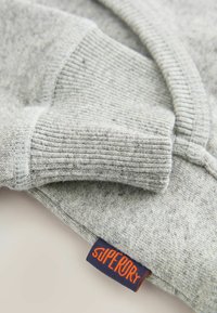 Felpa grigia con polsini a coste e una texture morbida. Include un'etichetta blu con testo arancione che recita "Superdry."