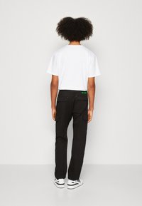 Persona con capelli ricci che indossa una maglietta bianca, pantaloni cargo neri con tasche e sneaker bianche, in piedi su uno sfondo uniforme.