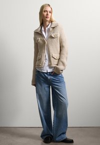 Beige knitted cardigan with large collar, two front pockets, and pearl buttons, paired with loose blue jeans and black shoes.