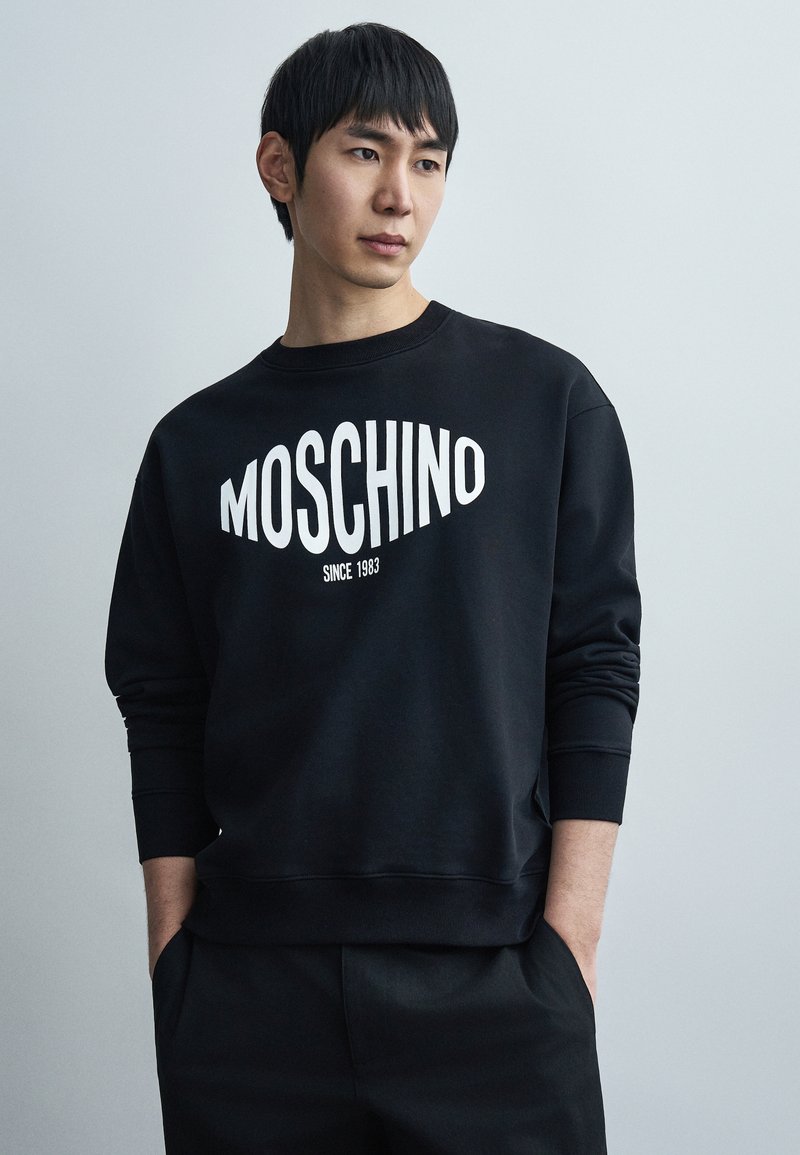 Μαύρο φούτερ με λευκό λογότυπο "MOSCHINO" και κείμενο "SINCE 1983". Διαθέτει ριμπ μανσέτες και λαιμόκοψη τύπου crew. Απαλή, ενιαία υφή υφάσματος.