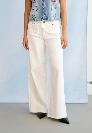 Kvinde iført hvide bukser med brede ben og snore i taljen, en blå denimvest med blomsterbroderi og sorte spidse sko.