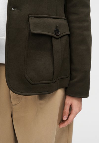 Olivo zelena blazer jakna z gumbom na poklopnem žepu, gladke teksture, strukturiranega dizajna in kontrastnim šivom, kombinirana z bež hlačami.