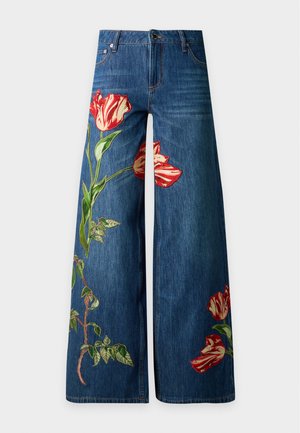 Femme portant un pull noir avec un motif de chapeau rouge et un jean bleu avec une grande broderie florale rouge, debout sur un fond blanc.