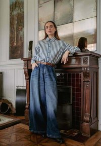 Una mujer vestida con una camisa a rayas azul y jeans de pierna ancha se apoya en una elaborada chimenea de madera en una habitación con un gran espejo y obras de arte enmarcadas.