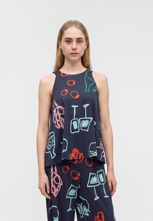 Jeune femme portant un top sans manches ample bleu marine et un pantalon avec des motifs abstraits colorés de verres à vin et de fruits sur un fond uni.