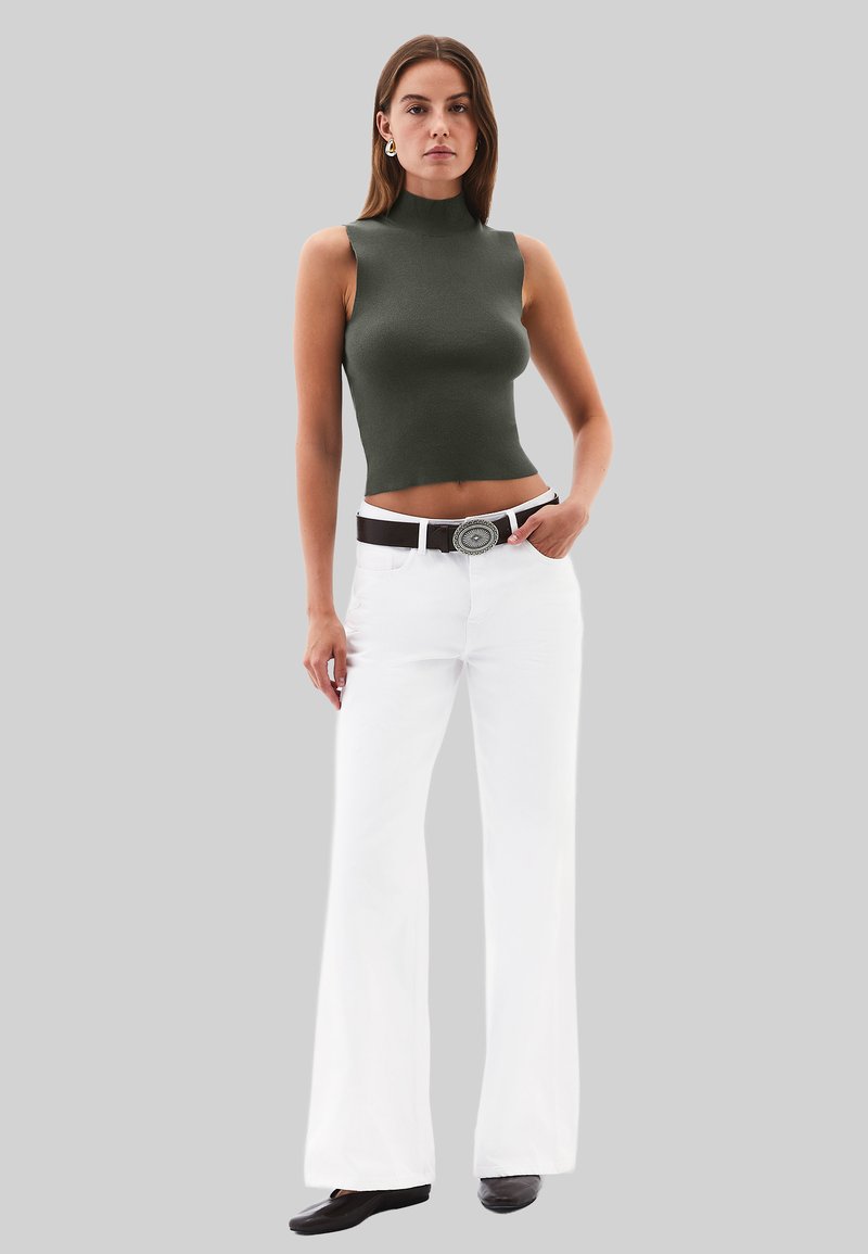 Top sin mangas ajustado de color verde oscuro, pantalones de pierna ancha blancos, cinturón negro con hebilla redonda de plata, textura suave, modelo de pie sobre un fondo gris.