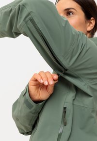 Grüne Outdoorjacke mit einer Reißverschlusstasche am Arm, strukturiertem Stoff und verstellbaren Handgelenkabschlüssen. Zeigt eine Nahaufnahme einer Hand, die den Reißverschluss benutzt.