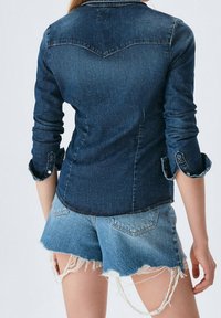 Kvinna iklädd en åtsittande mörk denimjacka med rullade ärmar och ljusblå slitna denimshorts, stående mot en enfärgad bakgrund.