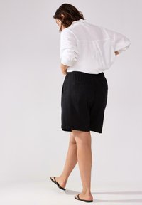 Pantaloni corti neri in lino con vita elastica e tasche laterali, abbinati a una camicia bianca dal taglio ampio. Il modello è scalzo e sta camminando.