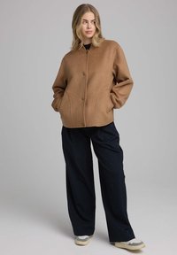 Bruine wolmix jas met een ronde kraag, voorknopen sluiting en zijzakken. Gecombineerd met losse zwarte broeken en off-white sneakers.