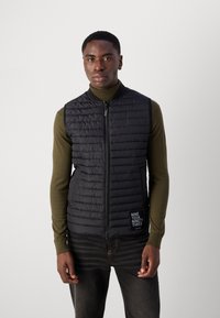 Zwart gewatteerd vest met een hoge kraag en ritssluiting aan de voorkant, met verticale stiksels en een logopatch aan de linkerkant. Draagt over een groene top.