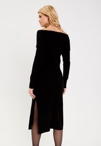 Robe noire à manches longues, décolletée épaules nues, avec un col nervuré et une fente sur le côté ; fabriquée en tissu doux avec une coupe ajustée.