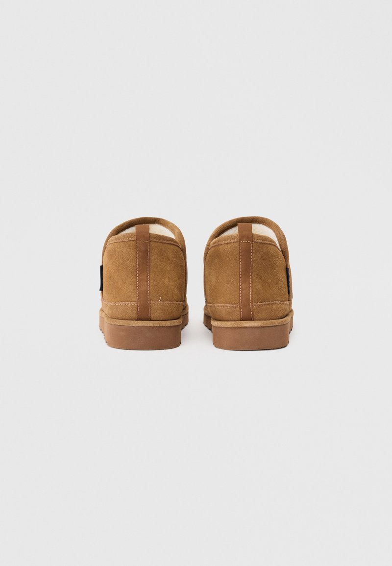 Chaussures en daim marron avec un dos arrondi, semelle plate en caoutchouc et accents en cuir. Doublure douce visible à l'ouverture.
