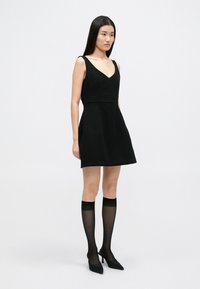 Μαύρο φόρεμα με V-λαιμόκοψη και εφαρμοστό κορμό, που ανοίγει σε φούστα. Διαθέτει τσέπες. Συνδυάζεται με ημίκοντες διαφανείς μαύρες κάλτσες και μυτερές γόβες.