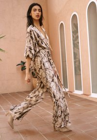 Mono fluido con estampado de serpiente en tonos beige y marrón. Tela ligera, pantalones anchos y cintura atada. Combinado con tacones transparentes.