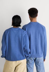 Dos personas con sudaderas azul oscuro de cuello redondo, mostrando la textura de la tela y las costuras de los hombros caídos, de pie una al lado de la otra frente a un fondo liso.
