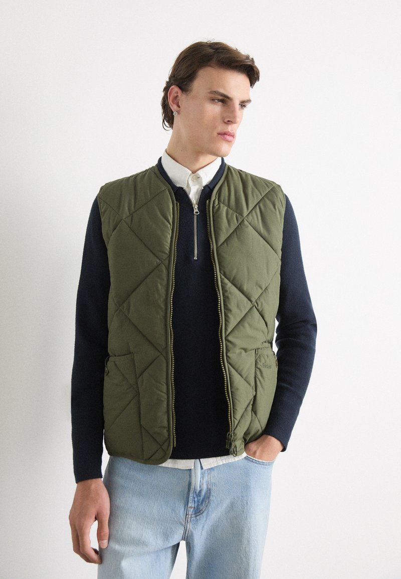 Gestepptes olivgrünes Weste mit einem Reißverschluss, zwei Taschen und einem gerundeten Kragen, getragen über einem navyblauen Pullover und hellblauen Jeans.