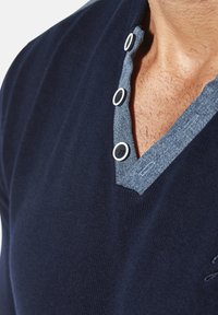 Marineblauer Strickpullover mit einem denimähnlichen Kragen. Verfügt über drei weiße Druckknöpfe und ein V-Ausschnitt-Design. Weiche Textur mit dezenten Nähten.