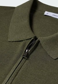 Gros plan d'un polo en maille vert olive avec un col replié et une fermeture éclair noire partiellement fermée.
