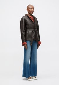 Καφέ δερμάτινο μπουφάν με υφή στον γιακά, ζωνάκι στη μέση και κόκκινη μπλούζα από κάτω. Μπλε φαρδιά τζιν και λευκά παπούτσια με μυτερή μύτη.