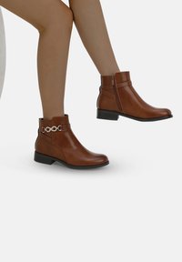 Botas de tobillo marrones de cuero suave, con una tira decorativa de cadena plateada y un cierre lateral para facilitar el uso. Tacón bajo y plano.