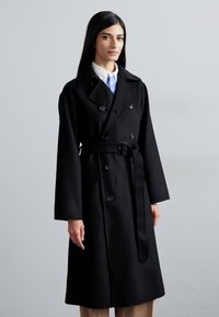 Μαύρο παλτό διπλής σειράς με ζώνη στη μέση, φαρδιά μανίκια και μεγάλα κουμπιά. Κατασκευασμένο από απαλό ύφασμα με κομψό σχέδιο.