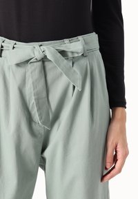 Calças de cintura alta verde-claro com cinto, frente plissada e bolsos laterais; feitas de um tecido suave com uma textura lisa.