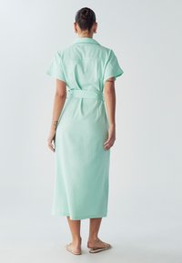 Mėtų žalios midi suknelė su trumpomis rankovėmis, diržu beliejamais juosmeniu, mygtukų apykakle ir lygiu audinio tekstūru, matoma iš nugaros.