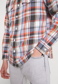 Camicia a quadri rossa, arancione, nera e blu con maniche lunghe, dettagli con bottoni e tasche sul petto, abbinata a jeans grigio chiaro.