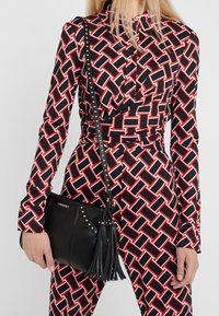 Borsa nera con borchie argentate e dettagli a frange; forma rettangolare; abbinata a un outfit con un motivo geometrico nei colori nero, rosso e bianco.