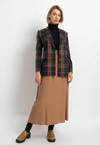 Karierte Blazer in Marineblau, Orange und Grau mit zwei Knöpfen, kombiniert mit einem braunen, fließenden Rock. Ein schwarzer Rollkragenpullover und Loafers runden das Outfit ab.