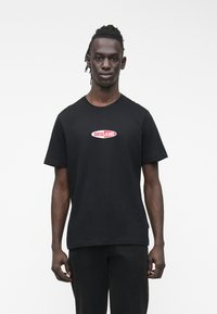 Melns kokvilnas T-krekls ar īsām piedurknēm un apaļu kaklu, uz krūtīm atrodas sarkans un balts "GUESS JEANS" logotips.