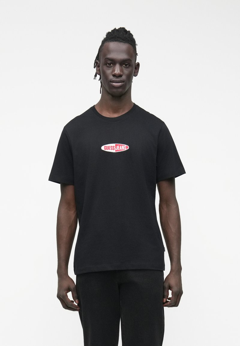 Melns kokvilnas T-krekls ar īsām piedurknēm un apaļu kaklu, uz krūtīm atrodas sarkans un balts "GUESS JEANS" logotips.