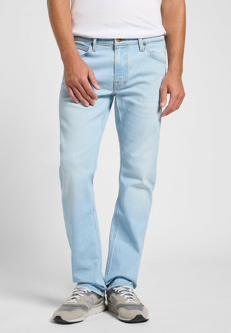 Jeans de mezclilla azul claro con corte recto, que presentan un clásico diseño de cinco bolsillos y una sutil textura desgastada. Llevados con zapatillas grises.