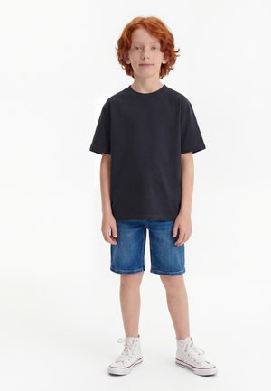 Băiat cu păr roșcat stând, purtând un tricou negru, pantaloni scurți din denim albastru și adidași albi cu gleznă înaltă pe un fundal alb.