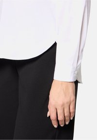 Main reposant contre un pantalon noir, portant une chemise blanche à manches longues avec des poignets boutonnés, sur un fond uni clair.