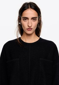 Mujer con cabello largo y oscuro que lleva pendientes de aro de plata y un suéter negro con dos bolsillos frontales, mirando directamente hacia adelante con una expresión neutral.