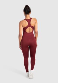 Bordeauxfärgad atletisk jumpsuit med racerback-design och cirkulär skärning, stretchigt tyg och åtsittande silhuett. Vita sneakers kompletterar looken.