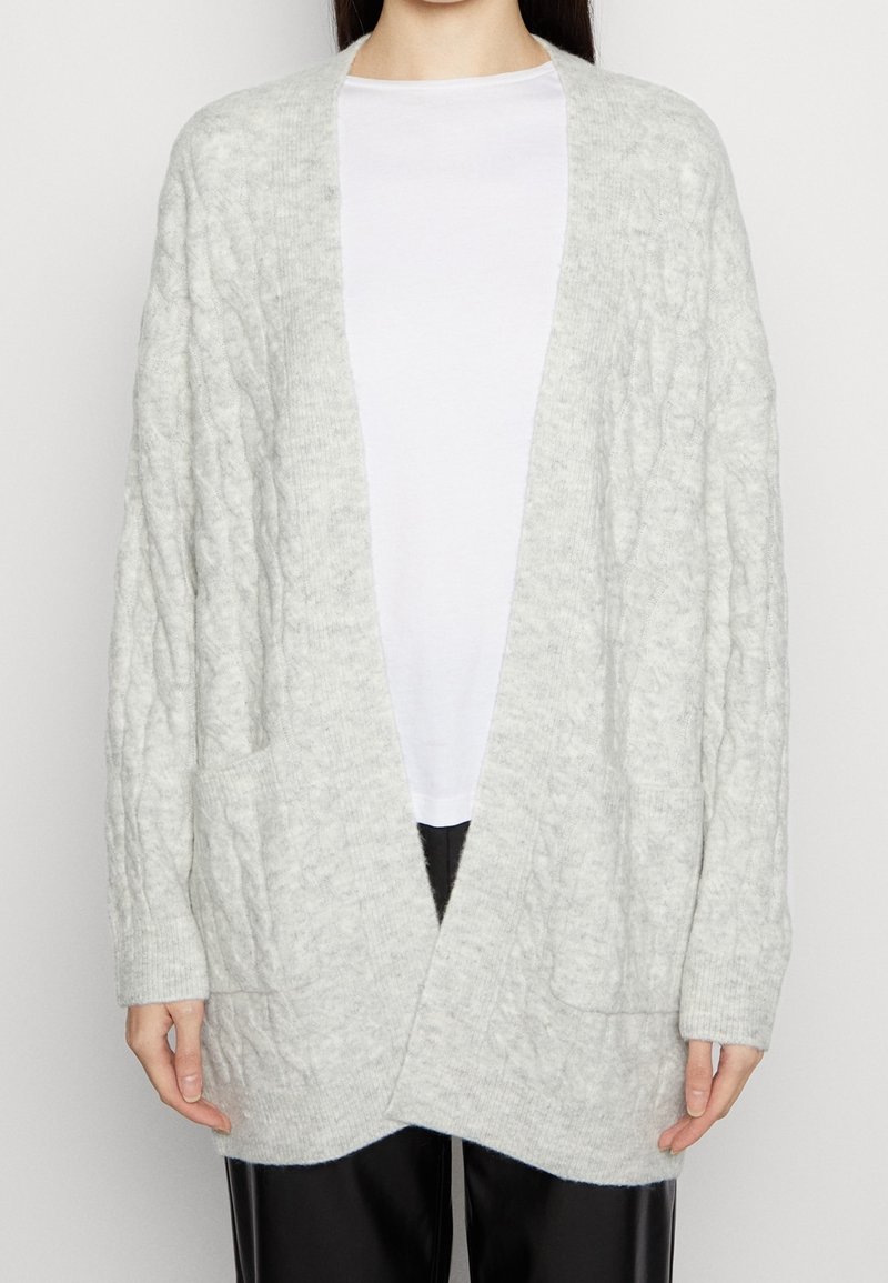 Femme portant un gilet épais en maille torsadée gris clair sur une chemise blanche avec un pantalon noir, debout devant un fond uni.