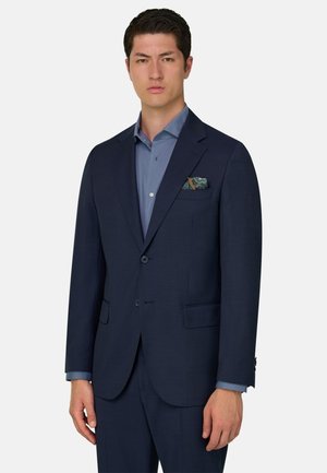 Hombre vistiendo un traje azul marino oscuro con un pañuelo de bolsillo con estampado y una camisa azul, sobre un fondo blanco liso.