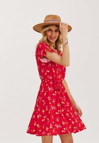 Czerwona sukienka w kwiaty z krótkimi falbaniastymi rękawami, z wcięciem w talii i rozkloszowaną spódnicą. Połączona z beżowym kapeluszem z ozdobną wstążką.