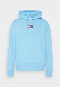 Ljusblå hoodie i mjuk bomull. Har en framficka, snörning i huvan och en liten märkespatch i rött och marinblått på bröstet.