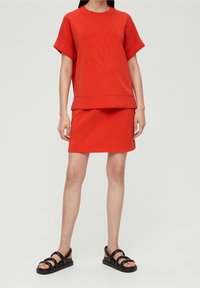Top corto de algodón rojo con diseño de hojas en relieve, combinado con una falda a juego. Sandalias negras con tiras completan el atuendo.